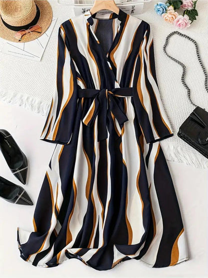 Burning Bones Abstract Print Surplice Neck Belted Dress styled in flatlay with navy, white, and caramel vertical stripes. Long-sleeve midi dress with wrap neckline and waist belt, shown alongside black pointed flats, straw hat, and decorative accessories on white background.